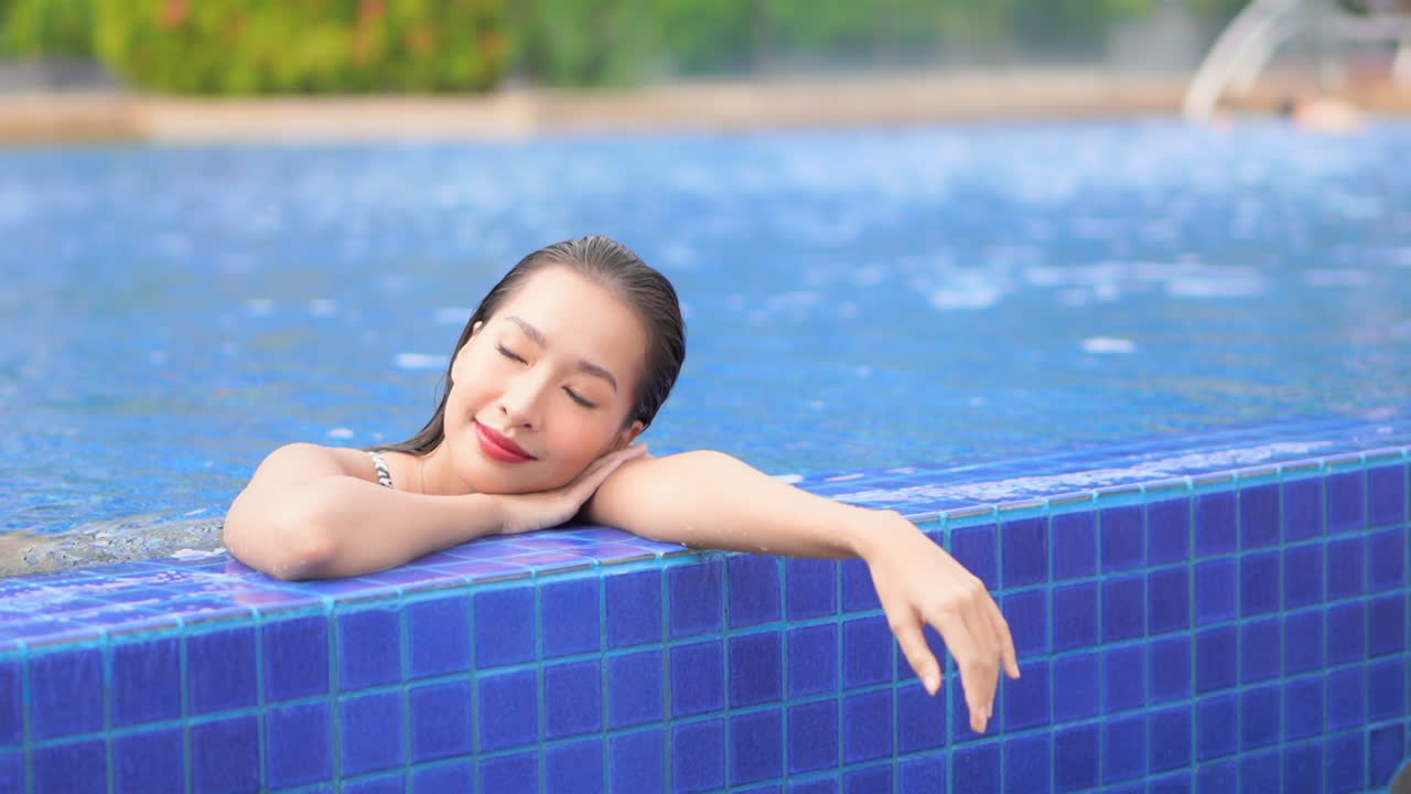 화려한 리조트 수영장에서 휴식을 취하는 동안 매력적인 젊은 여성이 팔에 머리를 얹고 눈을 감습니다.