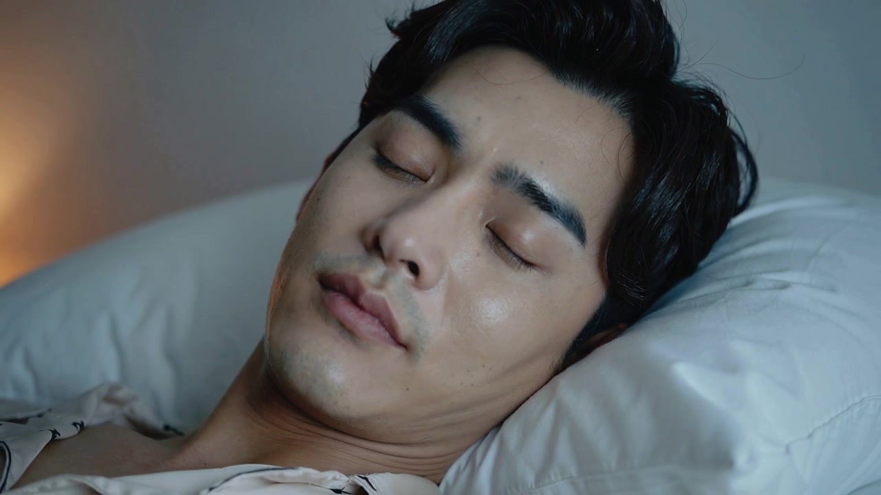 眠気を感じ、柄物の服を着た男性が目を閉じ、ランプの明かりの中で枕に頭を預けて眠りに落ちる様子