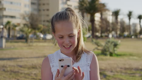 Retrato-Feliz-Niña-Rubia-Usando-Un-Teléfono-Inteligente-Disfrutando-Jugando-Juegos-Móviles-Sonriendo-Alegre-Satisfacción-En-El-Cálido-Parque-Al-Atardecer