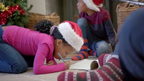 african american brother and sister in christmas hats drawing on floor in living room, slow motion