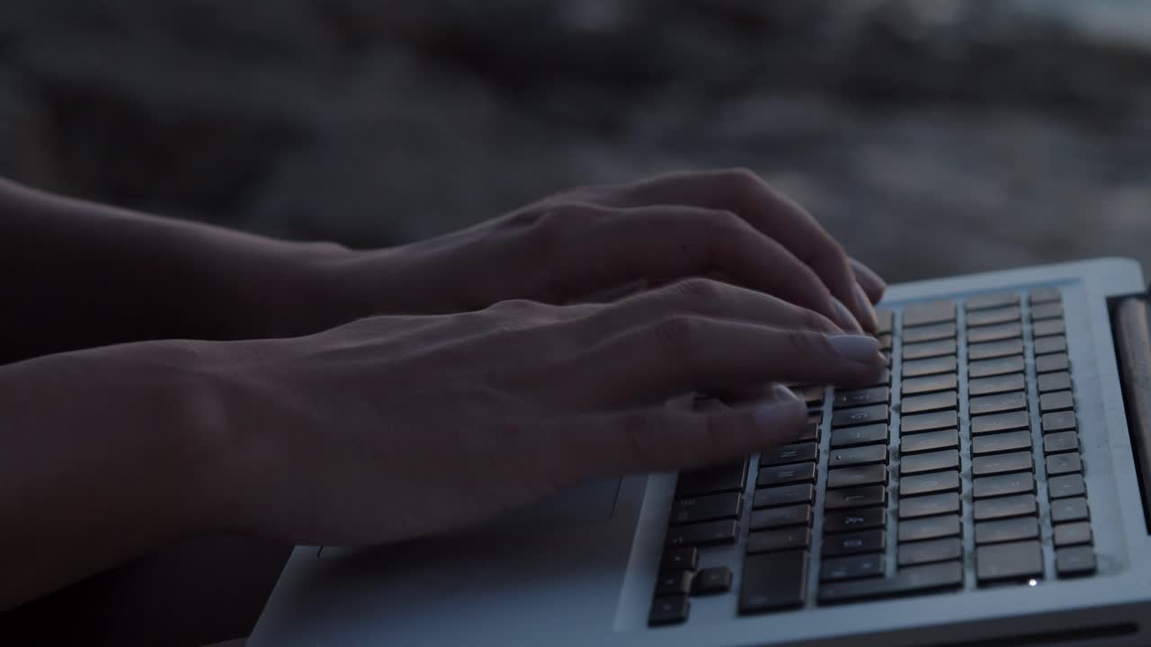 Piges hænder taster på laptop, siddende på en klippe ved havet, nærbillede.
