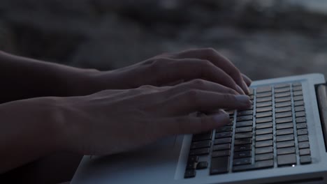 Girl-hands-typing-on-laptop-sitting-on-a-rock-by-the-sea,-close-up