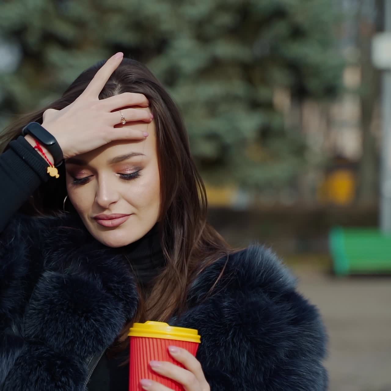Charming young woman in the park. Half-length portrait of a stylish girl in fur coat holds plastic cup of coffee on blur city background.