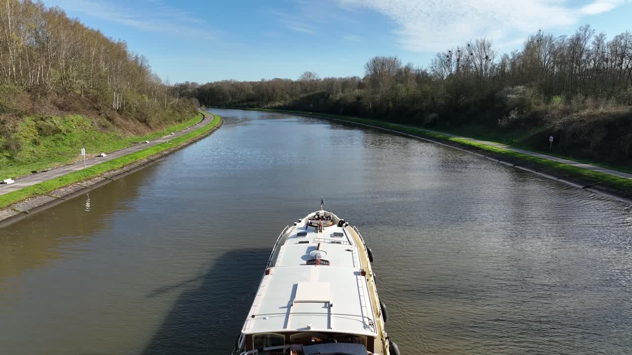 Static camera shot of boat advancing along canal in Brussels, Belgium, with countryside landscape and clear sky
