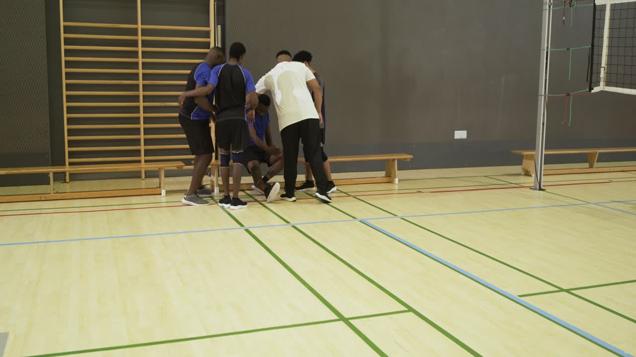 Diverse male volleyball players supporting limping teammate toward bench, crouching inspecting shoe