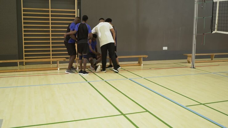 Diverse male volleyball players supporting limping teammate toward bench, crouching inspecting shoe