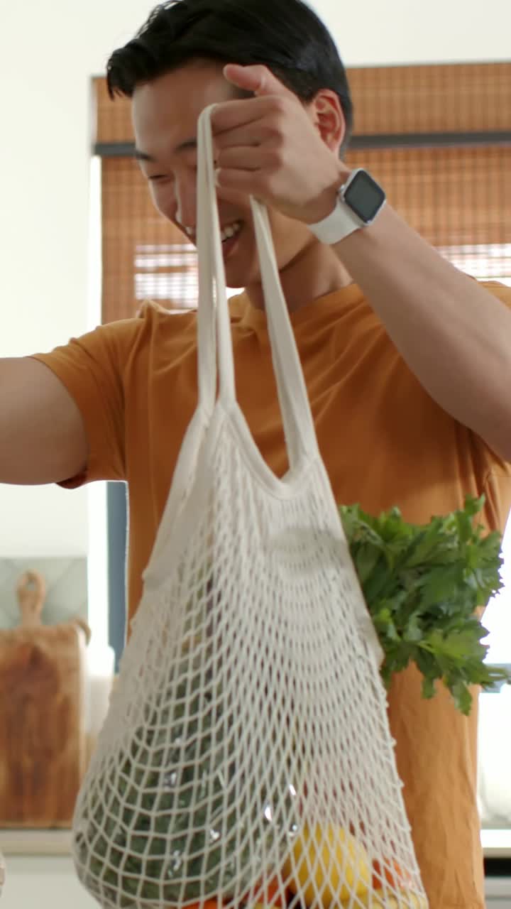 Vídeo vertical: Homem adulto de camiseta laranja erguendo uma sacola de rede na cozinha, dando um limão para uma criança.