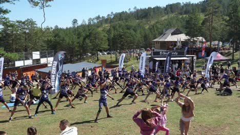 a large group of people engages in a fitness warm-up session outdoors, surrounded by trees and hills. participants stretch and exercise together, with banners and spectators in the background