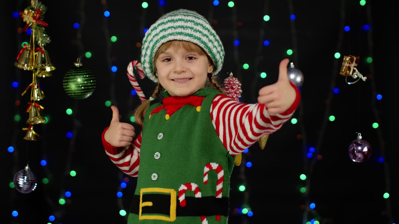 Náctiletá dívka v kostýmu vánočního elfa / pomocníka Santa Clause ukazuje palec nahoru na černém pozadí