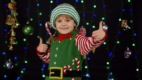 Teen-kid-girl-in-Christmas-elf-Santa-Claus-helper-costume-showing-thumbs-up-on-black-background