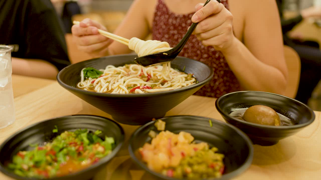 Woman lifts noodles from spicy soup with chopsticks, warm lighting, close-up, casual dining setting