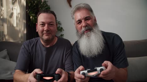 a happy brunette man with stubble in a gray t-shirt plays video games using a joystick with his boyfriend. an elderly man with gray hair and a lush beard he wins and rejoices. sitting on the sofa in a modern apartment in the cozy evening