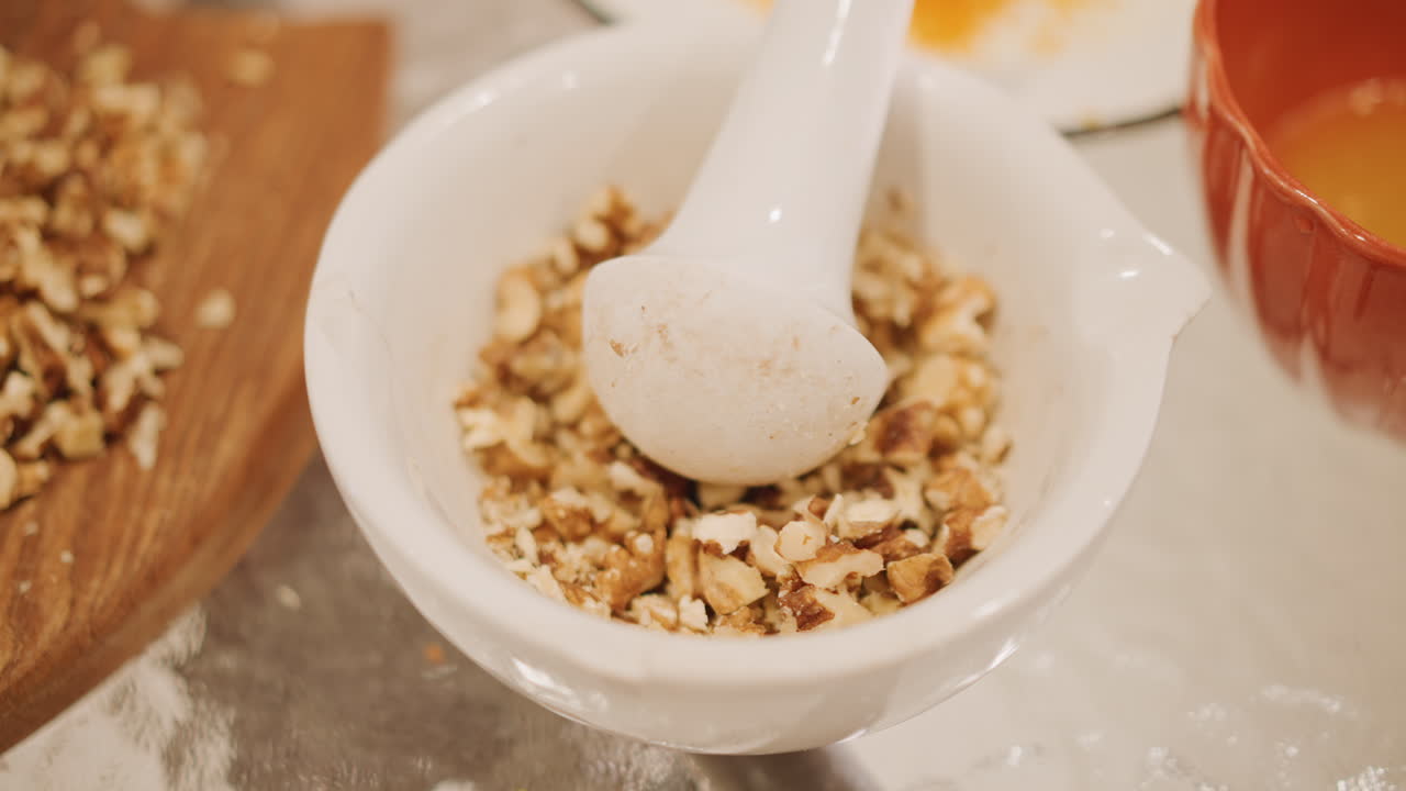 Preparazione di torta di zucca con noci tritate in un mortaio e pestello di porcellana. Un fornaio casalingo macina le noci pecan accanto a una ciotola di purea di zucca arancione. La calda superficie rustica della cucina è spolverata di farina. Primo piano sul ripieno croccante.