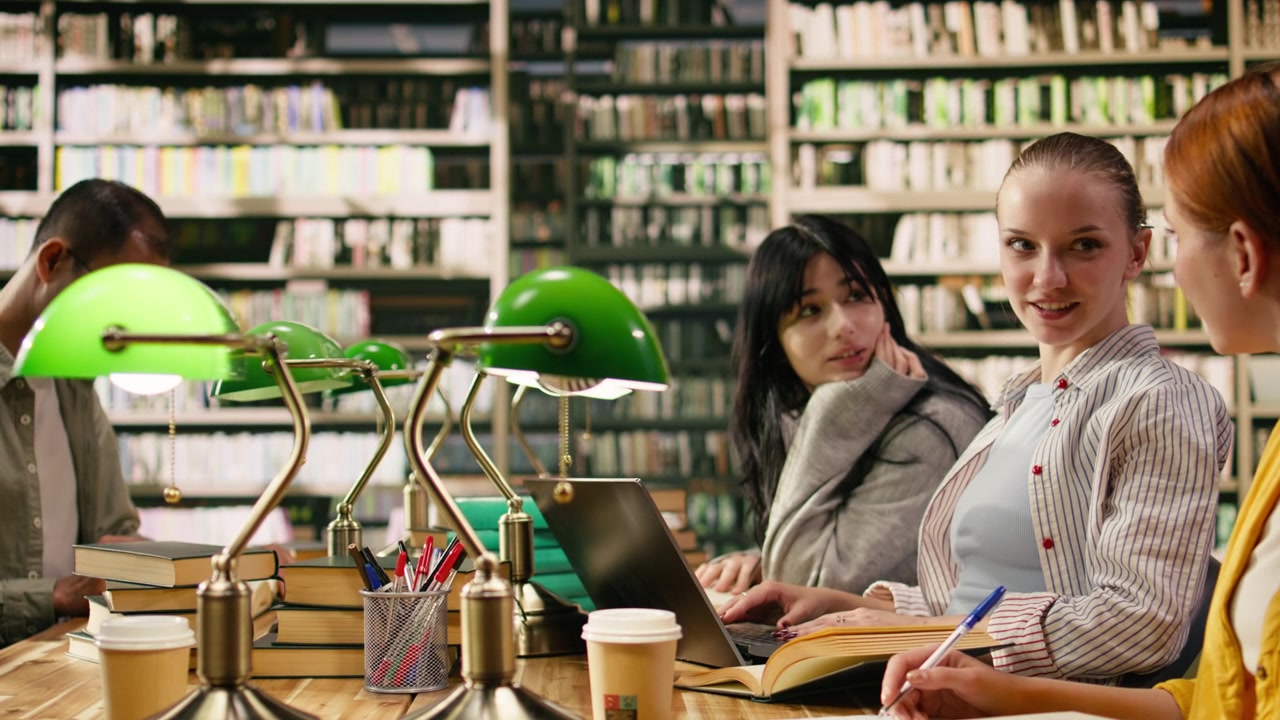 Studenti universitari discutono del lavoro del corso a un tavolo in biblioteca con libri aperti.