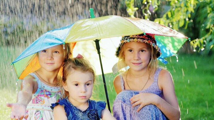 Tre bambine allegre e spensierate sotto l'ombrello dai colori dell'arcobaleno. C'è una pioggia calda e sono felici