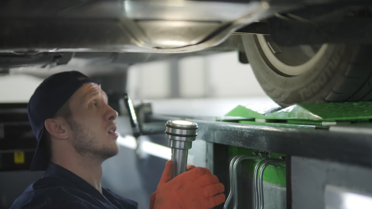 mecánico en uniforme reparando la parte inferior de un coche usando herramientas.