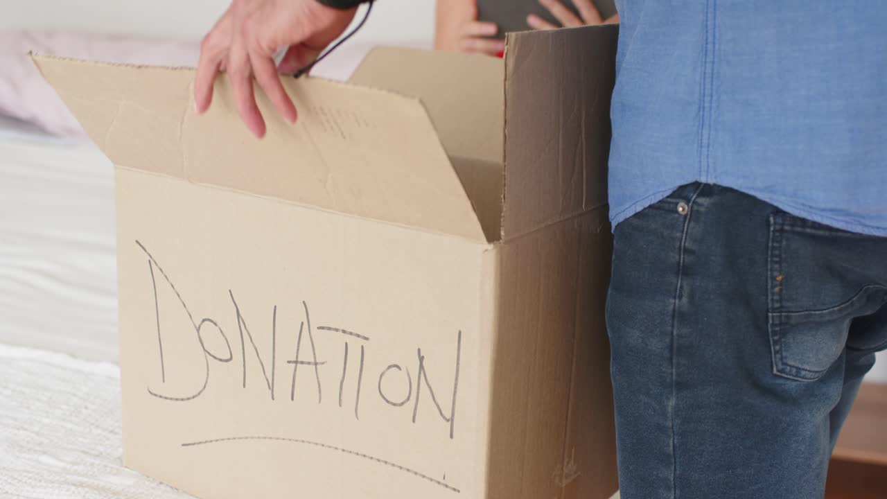 Couple packing donation box on bed, woman looking up and checking tablet while man folding flaps