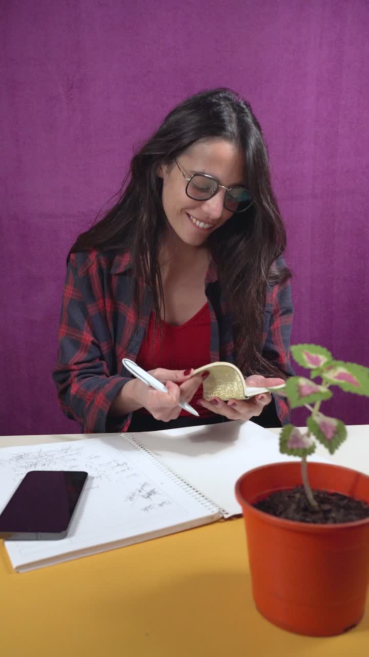 Uma jovem usando óculos escrevendo em uma mini agenda em um espaço de trabalho com uma planta em um vaso laranja, smartphone e um caderno grande
