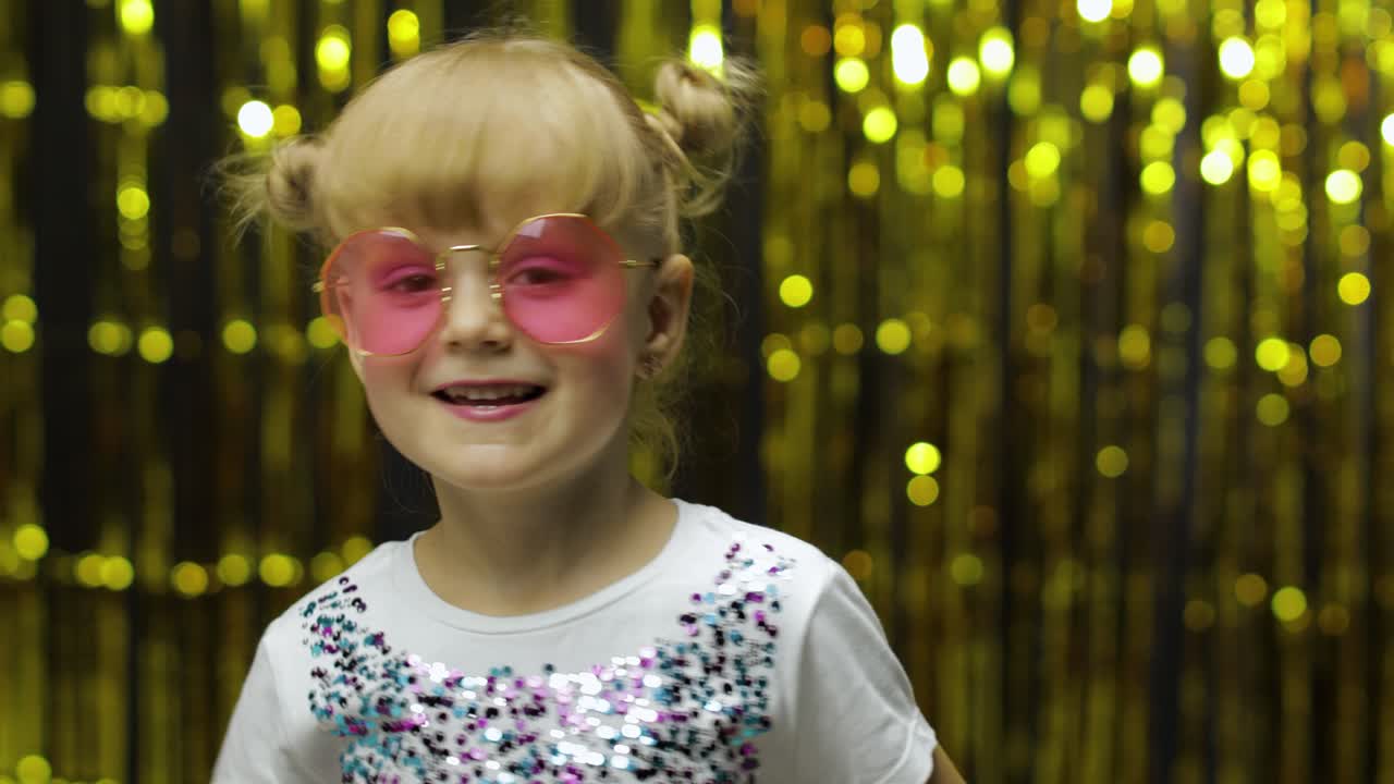 Child show thumbs up. Smiling, looking at camera. Girl posing on background with foil golden curtain