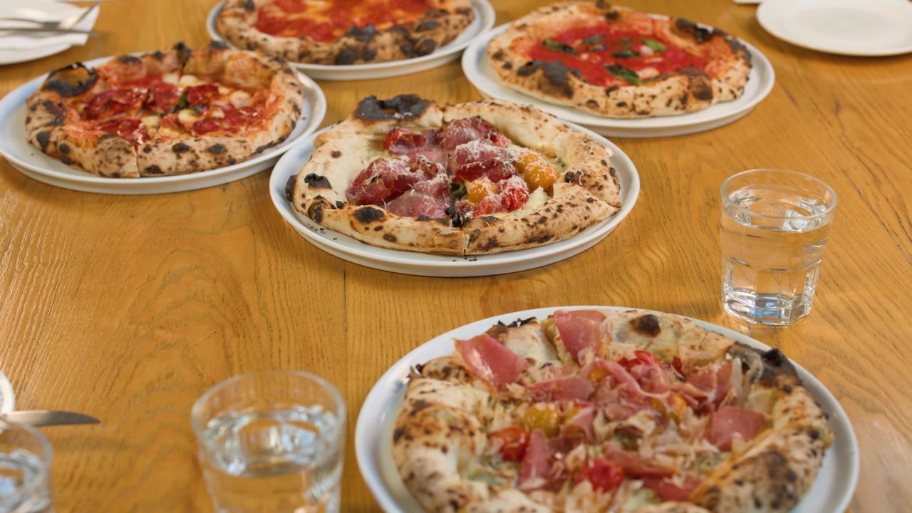 Una mano posiziona una pizza gourmet su un tavolo di legno pieno di piatti italiani.