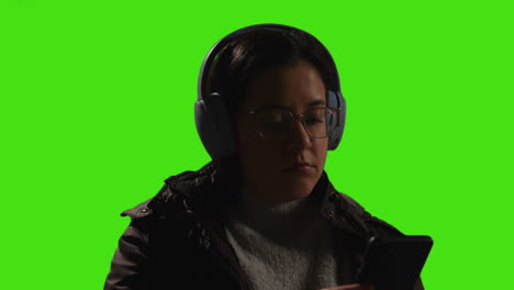 close up of woman wearing wireless headphones streaming music from mobile phone against green screen with low key lighting 1