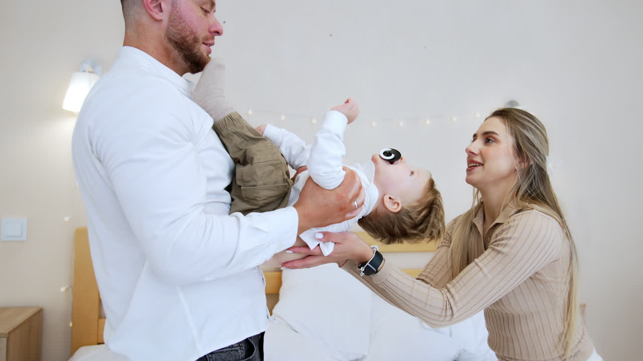 Little toddler boy tumbles from dad's arms into mum's. Happy joyful Caucasian family having fun in the bedroom together.