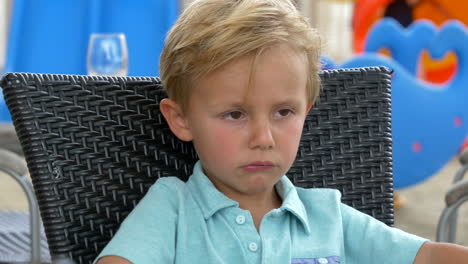 cerca de un niño pequeño y elegante rubio enojado caucásico sentado en una silla