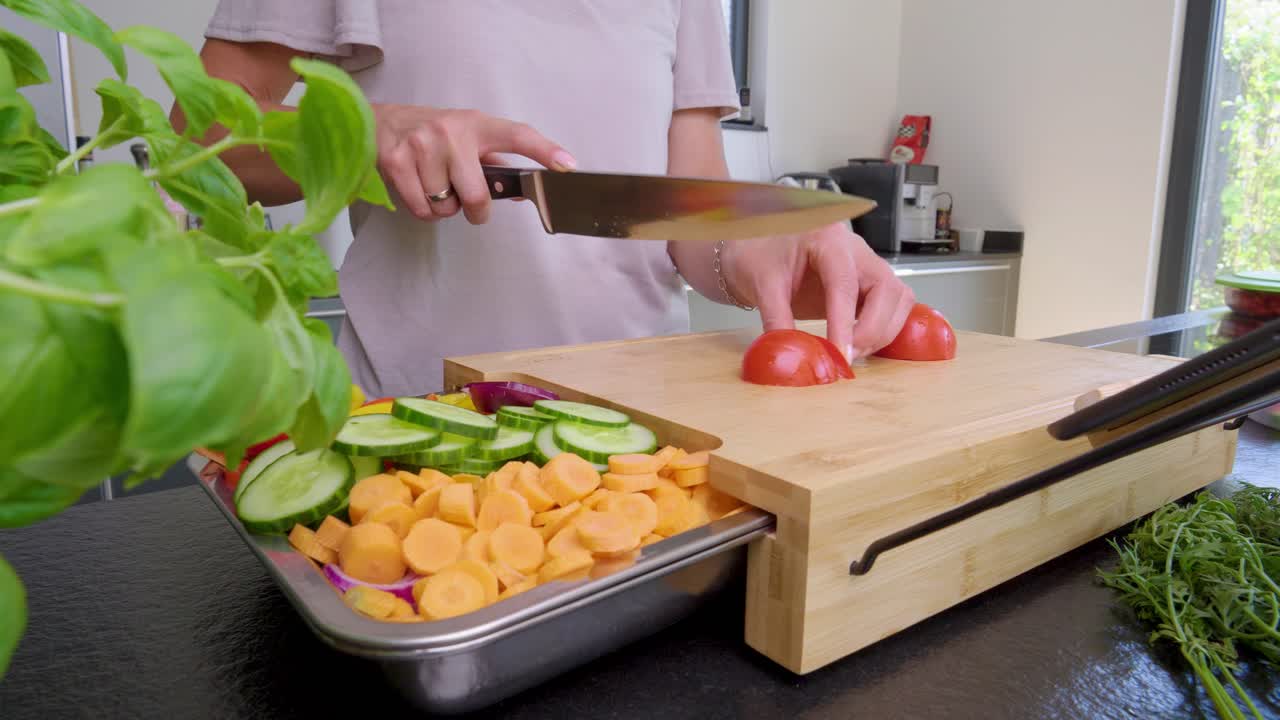 toma panorámica de una persona cortando un tomate en una tabla de cortar de madera en una cocina moderna y elegante con una bandeja de verduras preparadas