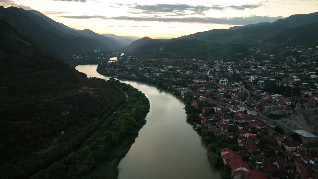 Ampia veduta aerea di una città tranquilla costruita lungo un fiume tortuoso, catturata durante il tramonto con montagne che incorniciano l'orizzonte e una luce calda che si riflette sull'acqua