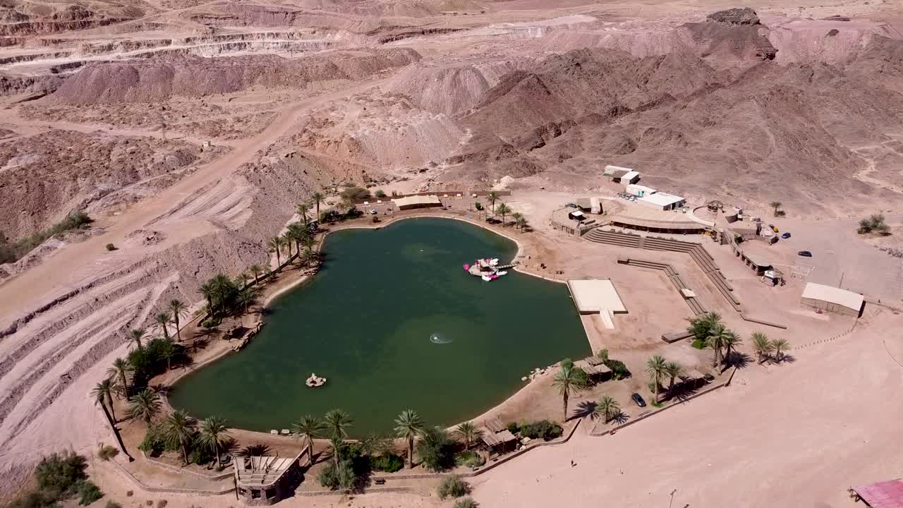 Ripresa aerea con drone dall'alto e in avanti di un'oasi artificiale in mezzo al deserto, circondata da palme, cespugli ed edifici