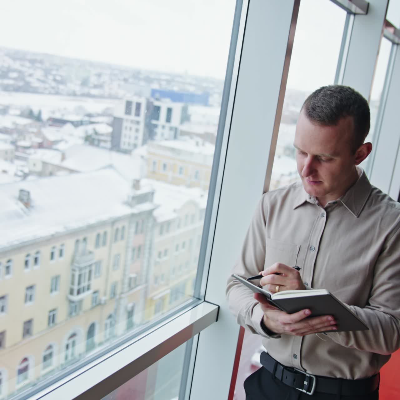 Caucasian man standing at the window and taking notes into notebook. Thoughtful male office worker at the backdrop of cityscape