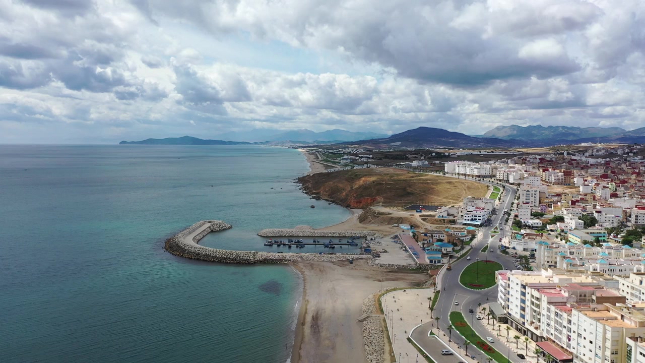 Ampia veduta aerea lungo la costa mediterranea di M'diq, che mostra il porto, i frangiflutti curvi, la strada costiera, il lungomare urbano e le montagne del Rif nel Marocco settentrionale, Nord Africa
