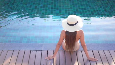 fashionable woman in sun hat relaxing sitting on the edge of swimming pool - back view