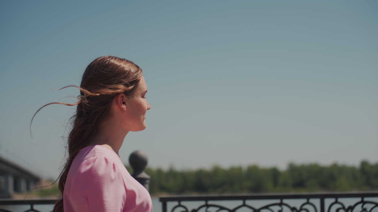 profilo di giovane donna vicino al fiume, capelli intrecciati che svolazzano nella brezza, camicetta rosa, ringhiera decorata e ponte in lontananza, luce solare soffusa e profondità di campo cinematografica, atmosfera contemplativa e quieta solitudine