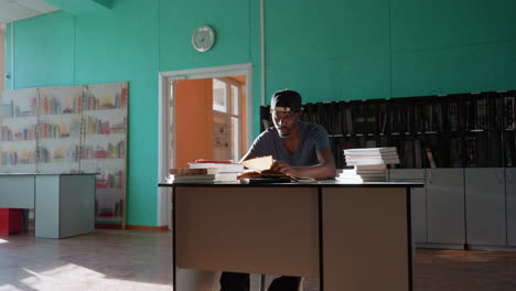Young man sits at desk in sunlit library, surrounded by stacked books, writing intently in notebook with pen, fully focused on study as light filters through windows