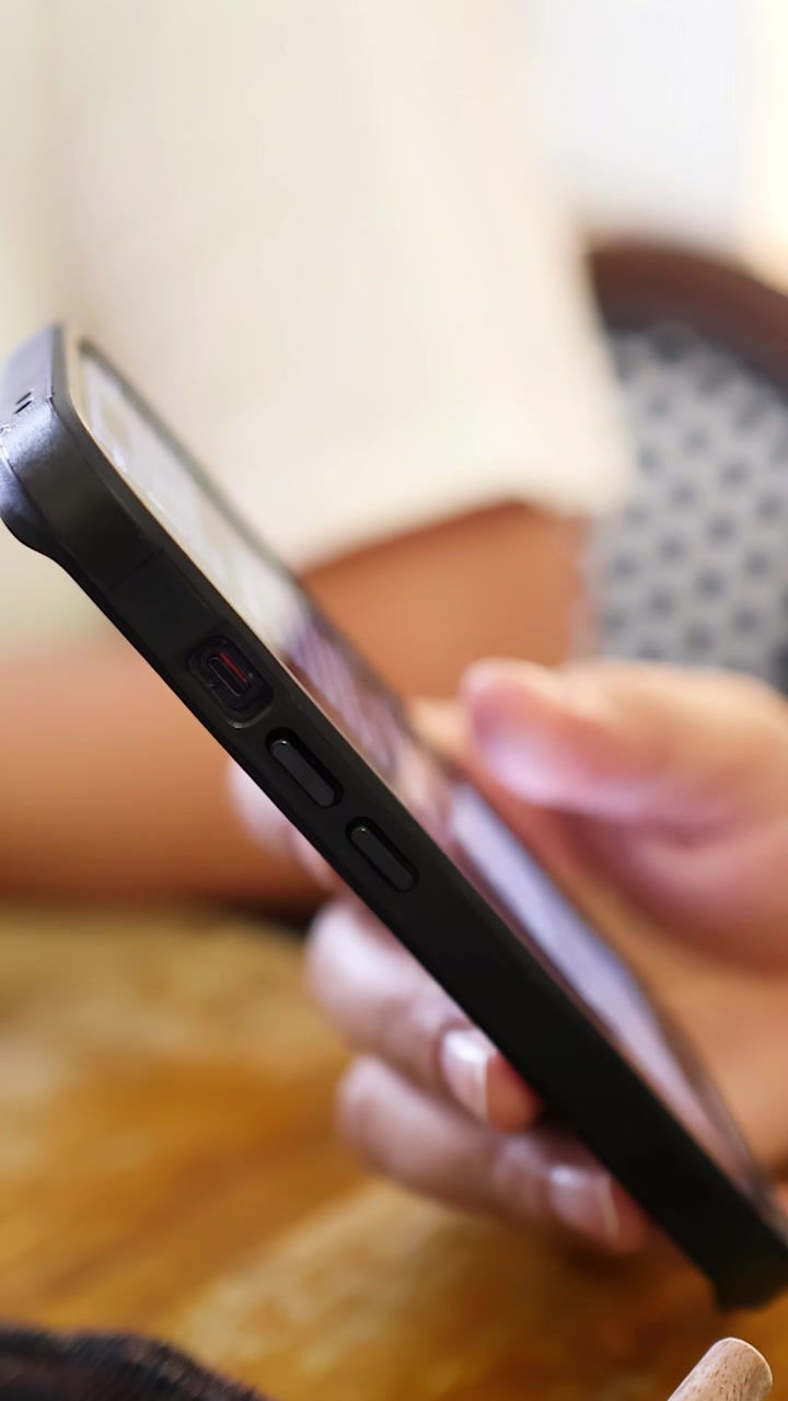Una persona interagisce con uno smartphone usando una mano, seduta a un tavolo di legno in un ambiente interno con luce soffusa. La telecamera rimane ferma, inquadratura laterale ravvicinata