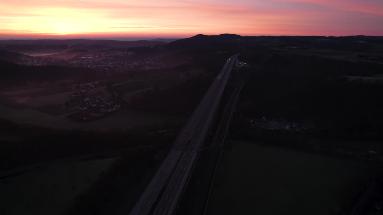 Tilting up drone footage showing morning traffic on a sprawling highway, bathed in the warm glow of a stunning orange-yellow sunrise, with fog lingering in the valley