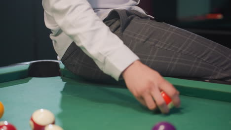 close-up of person in white shirt and grey trousers sitting on green pool table, rolling billiard ball. focus on lower body, stylish attire, and casual indoor ambiance. relaxed setting