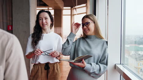 women working in the office chatting standing at the window. man sitting on desk in front of them puts on glasses and looks into the window.