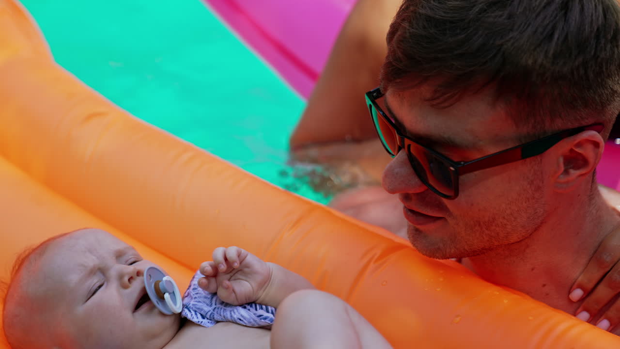 Beautiful blonde woman in black swimsuit swims on a mattress to her husband. Man is holding a mattress with a baby who cries. Mom tries to calm down the kid.