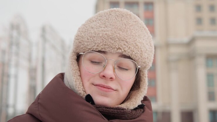 Profile view of elderly woman in knit headscarf, glasses, and warm coat walking through urban street under soft overcast daylight, with shallow focus, muted colors, and calm reflective mood