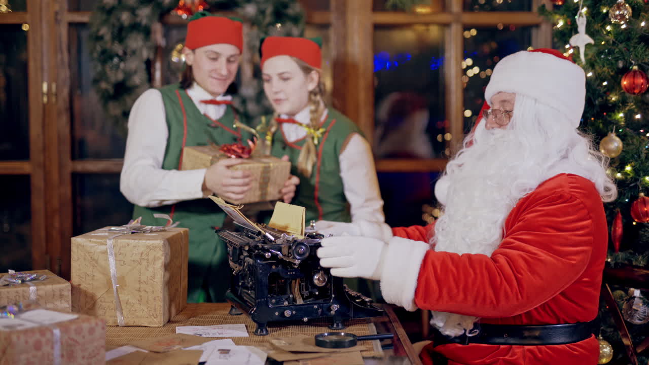 Santa and Elves with presents on Christmas decor indoor. Santa Claus is typing letter on an old typewriter at table. Happy elves in green costumes are dancing with present.