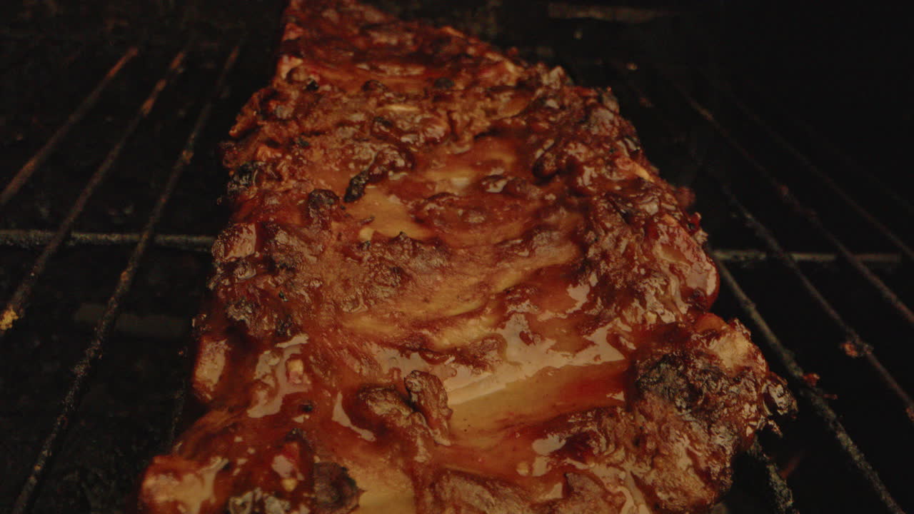Close up of spareribs being grilled on a barbecue. Smoke and flames rise from underneath the meat in slow motion