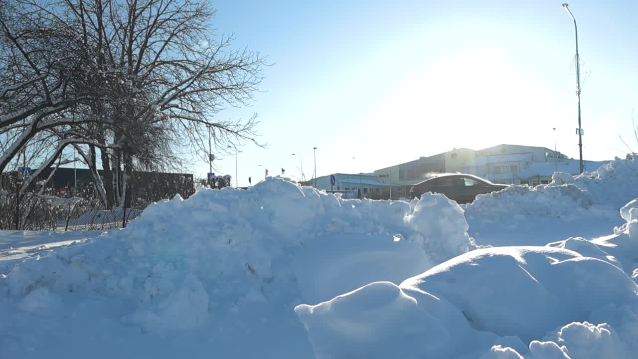 Low-angle panning view of snow-covered street in Laagri where thick white fluffy snow blankets the urban landscape, snowdrifts along the roadside while car drives by. Townscape emerges behind the snow