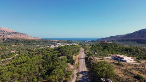 Sobre-La-Carretera-Y-El-Bosque-Verde-En-Rodas-Con-La-Playa-Al-Fondo-En-Grecia-Durante-El-Verano-Filmado-Con-El-Dron-En-4k