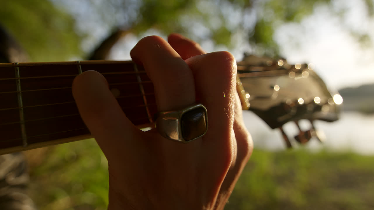Close-up of a person playing an acoustic guitar outdoors by a river.
