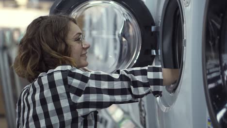 a young positive woman in a plaid shirt choosing washing machine in the shop of household appliances. look inside to the open door and smiling. side view. slow motion