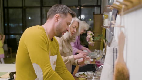 vista lateral de un hombre lavando los platos de la cena familiar en el fregadero de la cocina mientras habla con una mujer madura que está quitando los platos de la mesa