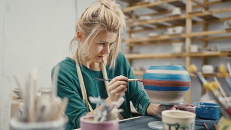Woman potter sitting at art studio and painting handmade ceramic pot, tracking shot, slow motion
