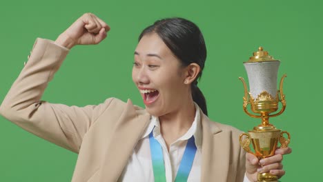 close up of asian business woman in a suit with a gold medal and trophy flexing her bicep and smiling to camera on green screen background in the studio